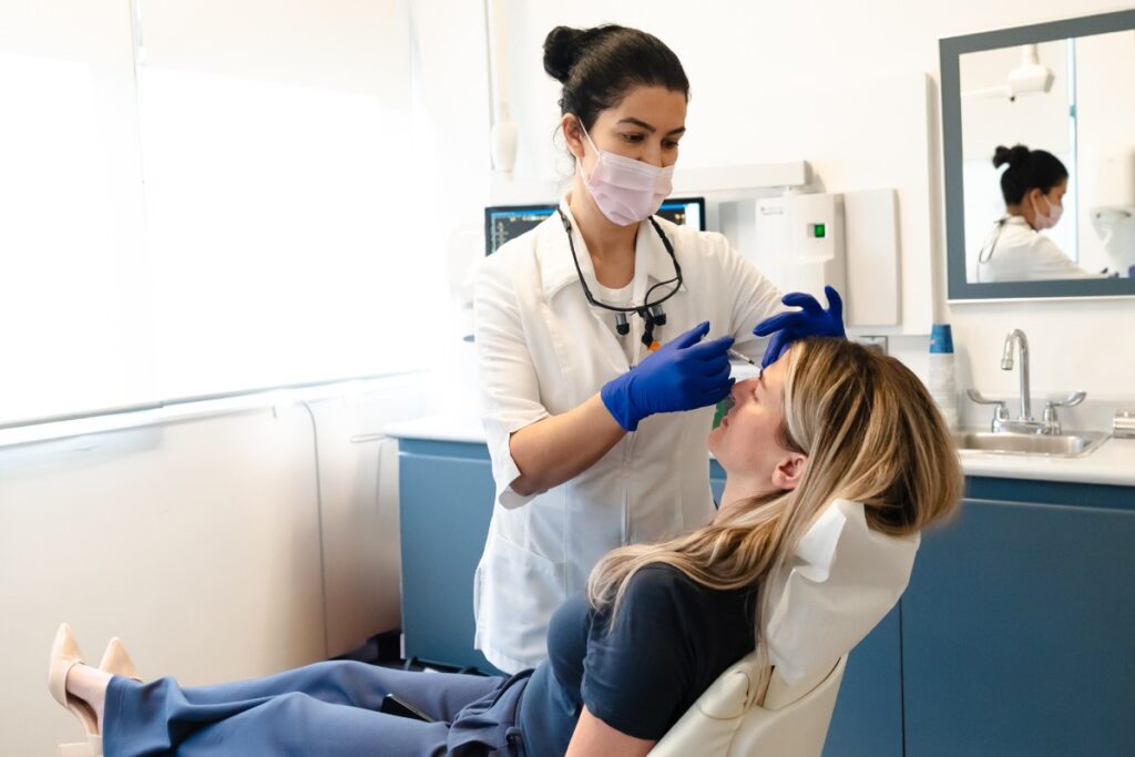 A dental hygienist wearing gloves and a mask is attending to a patient in a dental chair. The patient is reclining while the hygienist prepares to clean or examine their teeth. A mirror reflects their interaction in the background. The atmosphere is professional and caring.