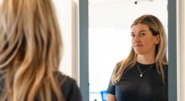 A woman with long blonde hair looks at her reflection in a mirror, displaying a thoughtful expression. The background is softly blurred, suggesting an indoor setting.