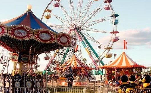 A vibrant carnival scene featuring a colorful carousel and a large Ferris wheel. Brightly painted amusement rides and a striped circus tent are visible, with people enjoying the festivities in a lively outdoor setting.