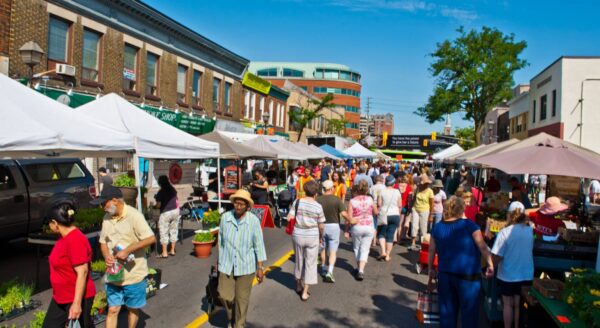 A bustling outdoor market setup with vendor tents, showcasing a variety of produce and goods. Shoppers, including diverse groups of people, browse the stalls under a clear blue sky, creating a lively community atmosphere.