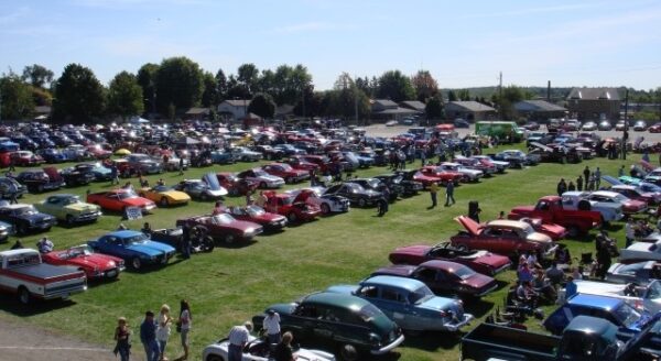 Aerial view of a large car show featuring numerous vintage and classic cars parked on a grassy field, with groups of people walking among the vehicles and enjoying the event under a clear blue sky.