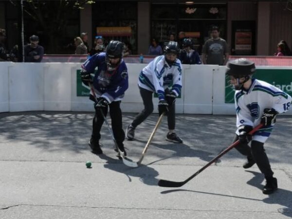Three players in hockey gear skate on a street hockey rink, competing for the puck. Two players in blue jerseys and one in white are actively engaging in the game, with onlookers in the background.