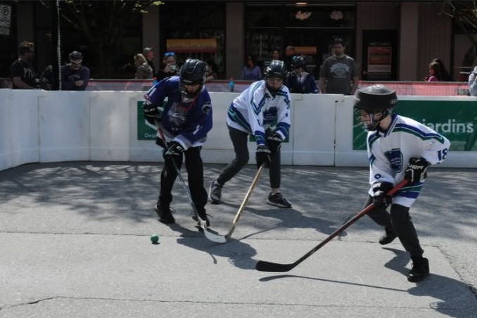 Three players in hockey gear skate on a street hockey rink, competing for the puck. Two players in blue jerseys and one in white are actively engaging in the game, with onlookers in the background.