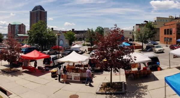 A vibrant outdoor market scene featuring various tents with red and white canopies, vendors, and visitors browsing. The setting includes trees and urban buildings under a clear blue sky.