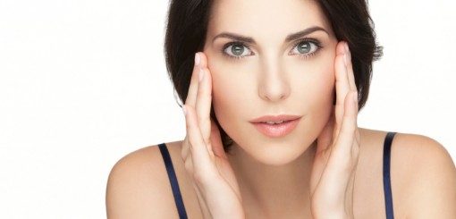A woman with dark hair and clear skin gently holds her face with both hands, looking directly at the camera with a neutral expression. The background is plain, emphasizing her features.