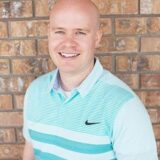 A smiling bald man is wearing a light blue and white striped polo shirt standing in front of a brick wall.