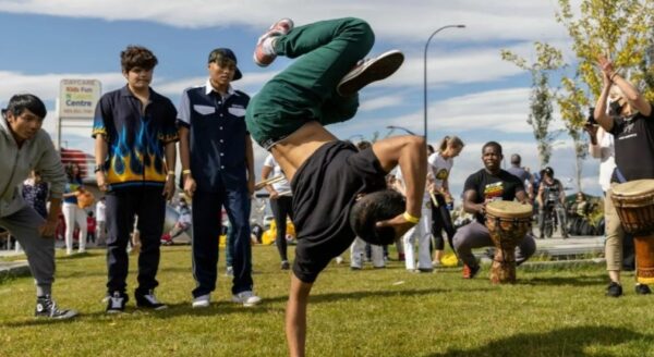 A breakdancer performs a skillful handstand on grass in front of an engaged crowd. Nearby, spectators cheer and play drums, contributing to the energetic atmosphere of the outdoor event. The scene showcases vibrant movement and cultural expression.