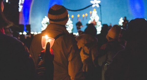 A crowd of people in winter clothing, holding candles, gathered at a festive event with illuminated trees and decorations in the background. A stage with a star-shaped decoration is visible, creating a warm, joyful atmosphere.
