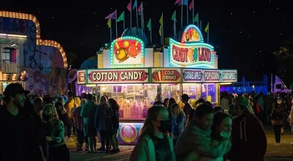 A brightly lit cotton candy stand is bustling with people at a nighttime fair, surrounded by colorful flags and amusement rides, creating a lively, festive atmosphere.