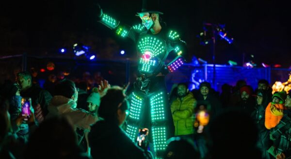 A performer in an illuminated costume made of colorful LED lights stands on stilts, engaging with a crowd during a nighttime festival. The background is filled with various festival-goers, creating a lively atmosphere.