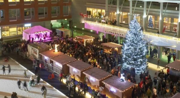A festive outdoor scene featuring a large decorated Christmas tree surrounded by a bustling crowd. There are wooden market stalls offering holiday goods, with colorful lights illuminating the area at night. People skate on an ice rink nearby, enjoying the holiday atmosphere.