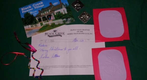 A collection of items arranged on a green surface, including a postcard from Black Creek Pioneer Village, a letter, two colorful blank cards, and decorative ribbon. The postcard features a historic building, while the letter expresses greetings.