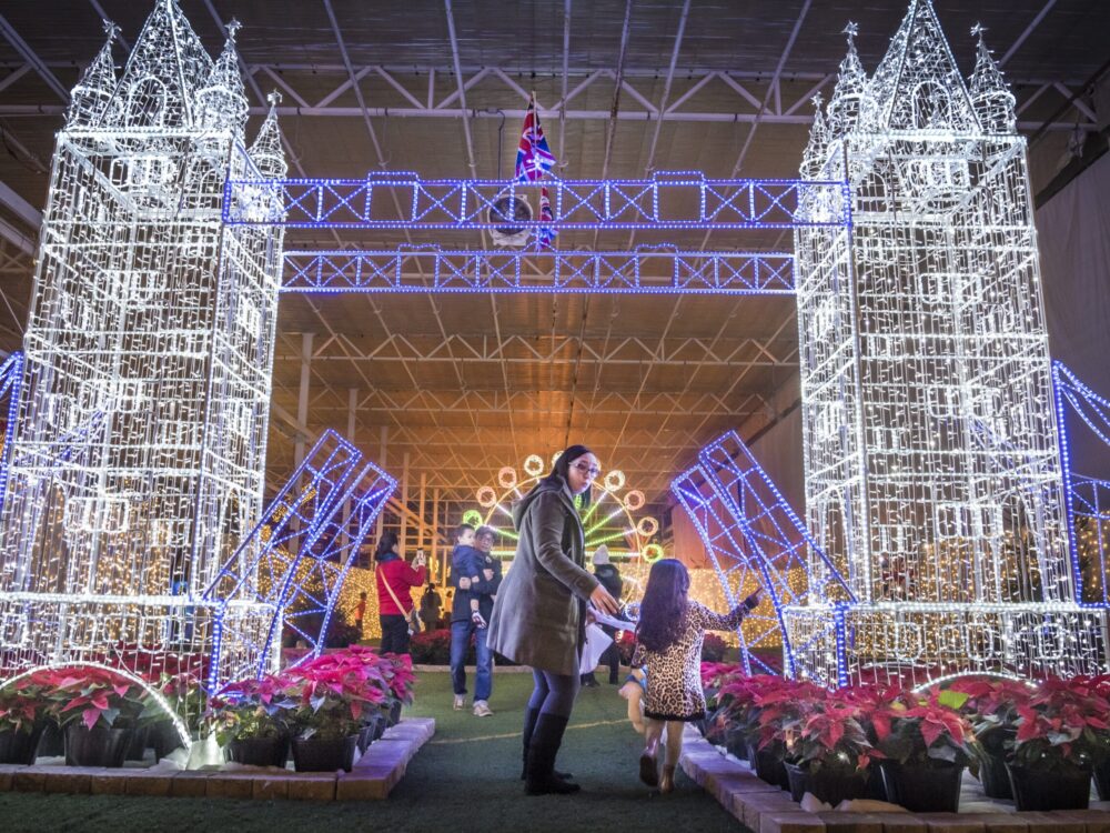 A woman and a child play under a dazzling, illuminated arch resembling a landmark, with vibrant flowers surrounding them. The scene is festive and magical, exuding a warm holiday atmosphere.