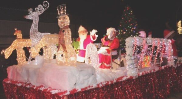 A festive parade float decorated with Christmas lights features Santa Claus, a reindeer, a nutcracker, and a snowy backdrop. The float is adorned with red glittery material and a Christmas tree, creating a cheerful holiday scene at night.