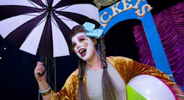 A cheerful clown in vibrant attire, holding a striped umbrella and a beach ball, smiles joyfully. The backdrop features colorful decorations, contributing to a festive atmosphere.