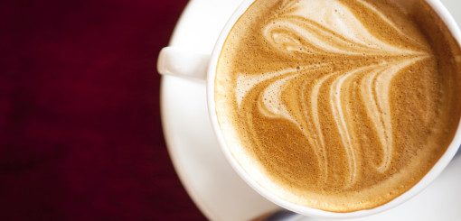 A close-up view of a latte in a white cup, showcasing intricate foam art resembling a leaf pattern, placed on a saucer against a deep red background.