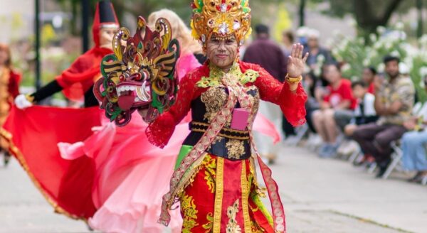 A performer wearing elaborate traditional attire smiles while dancing in a vibrant cultural festival. The colorful outfit features intricate decorations, and other dancers in the background wear flowing costumes. Spectators watch and enjoy the lively celebration.