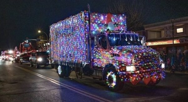 A brightly lit truck adorned with colorful holiday lights drives down a street at night, surrounded by festive decorations and other vehicles in a parade setting.