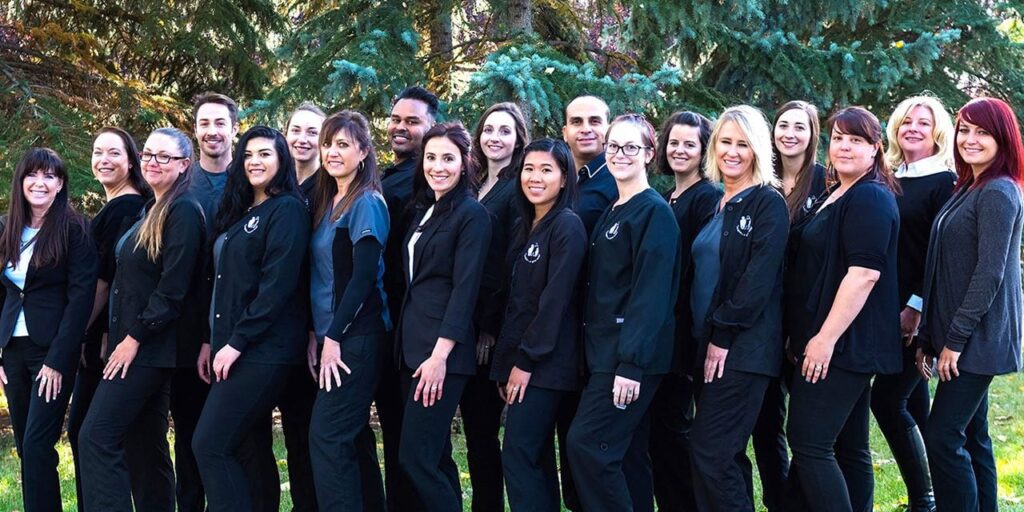 A group of nineteen people, all wearing matching dark outfits, stand in a line outdoors with trees in the background, smiling at the camera.