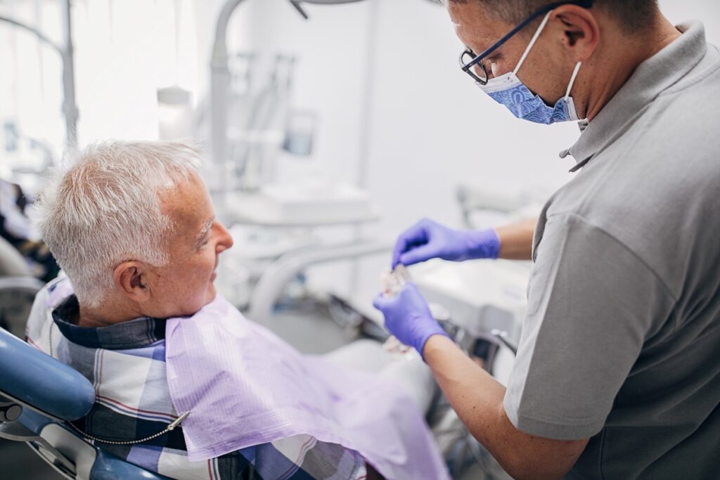 A dentist in a gray shirt wearing gloves interacts with an elderly patient wearing a dental bib in a modern dental office. The patient looks relaxed as the dentist prepares for a procedure, demonstrating a caring atmosphere in dental care.