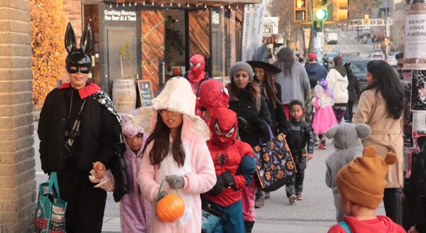 A group of children and adults in various costumes, including superheroes and a bunny, stroll along a city street during a festive event. The scene features colorful outfits and Halloween-themed decorations as they walk past shops and traffic lights.