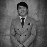 A smiling man in a double-breasted gray suit with a white shirt and patterned tie stands confidently against a textured concrete wall, exuding a professional demeanor.