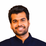 A smiling man with short black hair and a well-groomed beard wearing a dark blue traditional outfit against a white background.