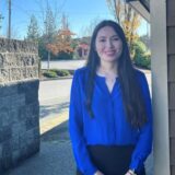 A woman with long dark hair smiles while standing outside, wearing a blue blouse and black skirt. The background features trees with autumn leaves and a clear blue sky.