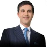 A well-dressed man with dark hair smiles confidently while wearing a tailored dark suit and a light blue tie. The background is plain white, emphasizing his professional appearance.