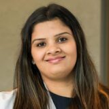 A smiling woman with long, dark hair, wearing a white coat over a black top, poses for the camera in a professional setting.