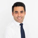 A professional portrait of a smiling man with dark hair, wearing a white shirt and a navy blue tie against a plain white background.