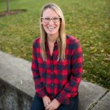 A smiling woman with long blonde hair sits on a stone ledge outdoors, wearing a red and black plaid shirt and glasses, surrounded by grass and fallen leaves.