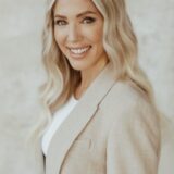 A smiling woman with long, wavy blonde hair wears a light-colored blazer over a white top. She stands against a neutral background, exuding a professional and approachable demeanor.
