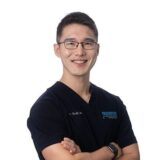 A smiling male medical professional wearing glasses and a dark scrub top stands with his arms crossed, conveying a sense of approachability and confidence.