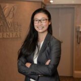 A young woman with long black hair and glasses stands confidently with her arms crossed, wearing a gray blazer over a white shirt. The background features signage for Bayview Village Dental.