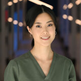 A smiling dental professional wearing a green scrub top poses confidently in a well-lit hallway, conveying warmth and approachability.