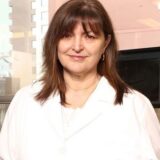 A woman with long brown hair, wearing a white lab coat, stands in a well-lit indoor setting with a window in the background. She smiles confidently, exuding a professional demeanor.