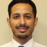 A professional headshot of a man with short dark hair and a slight beard, wearing a white shirt and a patterned tie, against a plain light-colored background.
