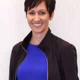 A smiling woman with short dark hair is wearing a black jacket over a blue shirt. She stands against a plain light background, exuding a friendly and approachable demeanor.