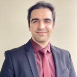 A smiling man in a suit and tie stands against a neutral background, exuding professionalism and confidence.