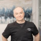 A smiling male dentist in a black medical uniform stands confidently in an office setting, with abstract art in the background.
