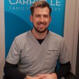 A smiling man wearing scrubs sits in front of a sign for "Carryville Family Dentistry." He has short hair and is positioned with hands resting on his knees, conveying a friendly and professional demeanor.