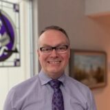 A smiling man with glasses and short hair, wearing a light purple shirt and patterned tie, stands in an indoor setting with a colorful stained glass window and a framed painting in the background.