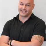 A smiling man with a shaved head and tattoos on his right arm stands with his arms crossed, wearing a black polo shirt against a light background.