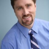A smiling man with brown hair and a goatee wears a blue dress shirt and a striped tie. He is posed against a light blue background, conveying a professional and approachable demeanor.