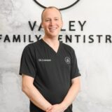 A smiling dentist in a black scrub stands in front of a wall sign that reads "Valley Family Dentistry." The background features a modern, minimalist design.