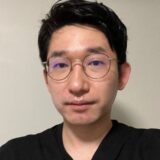 A young man with short black hair and wearing round glasses poses for a selfie. He is dressed in a black shirt and has a neutral expression, with a plain background behind him.