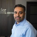 A smiling man with a beard, wearing a light blue shirt, stands in an office setting. The background features a sign that reads "Axler Dental Centre."