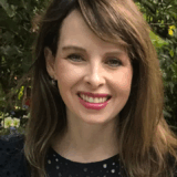 A woman with long, brown hair smiles while wearing a navy blue, polka-dot blouse. She is outdoors, surrounded by greenery.