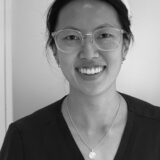 A smiling individual wearing glasses and a dark shirt stands against a light background. The person's hair is pulled back, and they appear to be in a professional setting, conveying approachability and confidence.